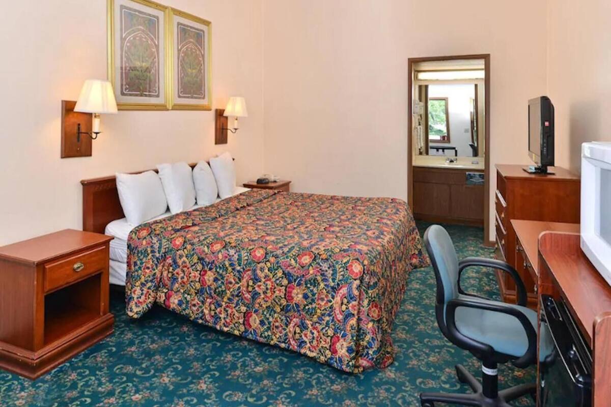 A hotel room with a bed, a desk, a chair, a TV, and two wall-mounted lamps.