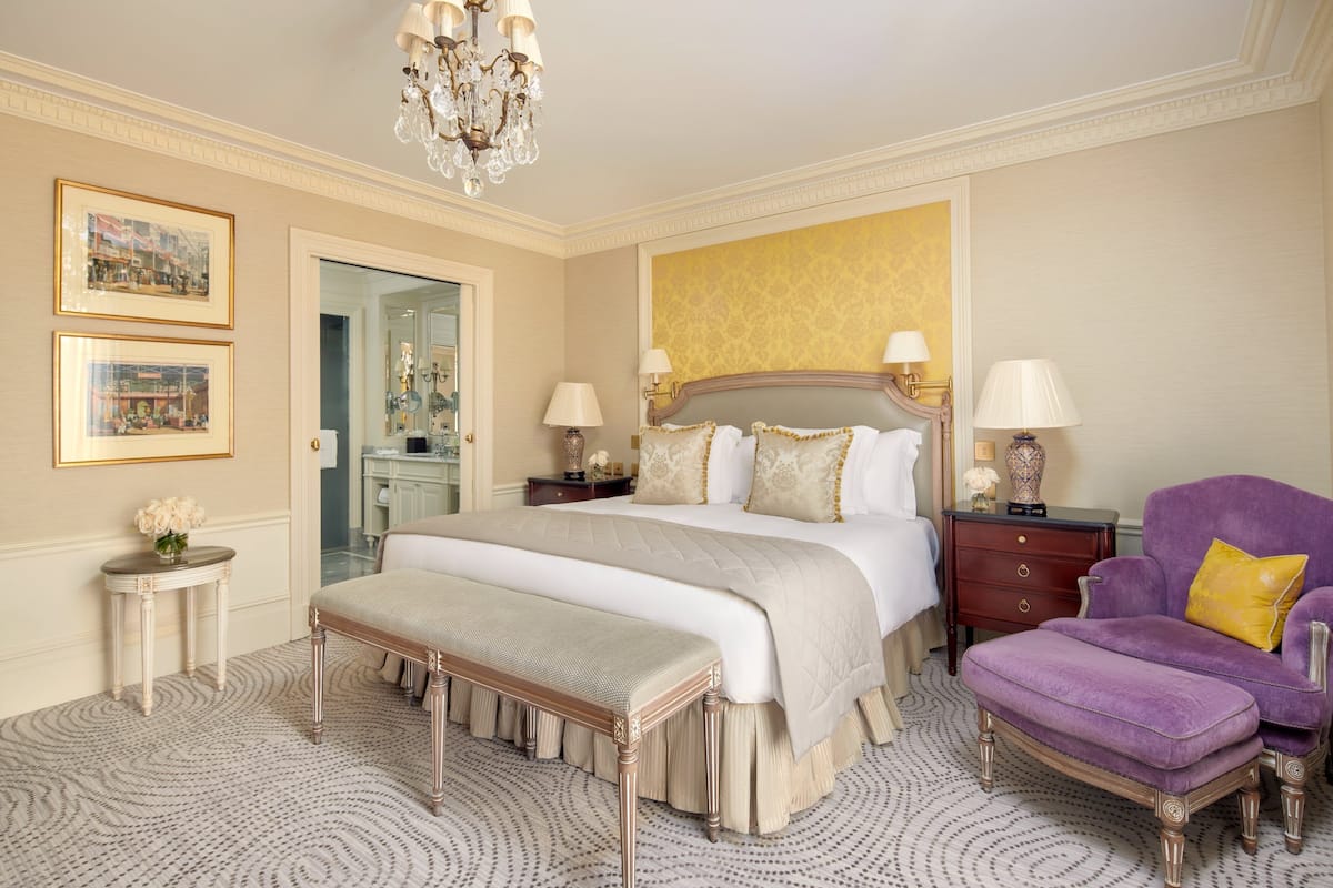 A spacious bedroom with a large bed, a bench, a purple armchair, and a chandelier.