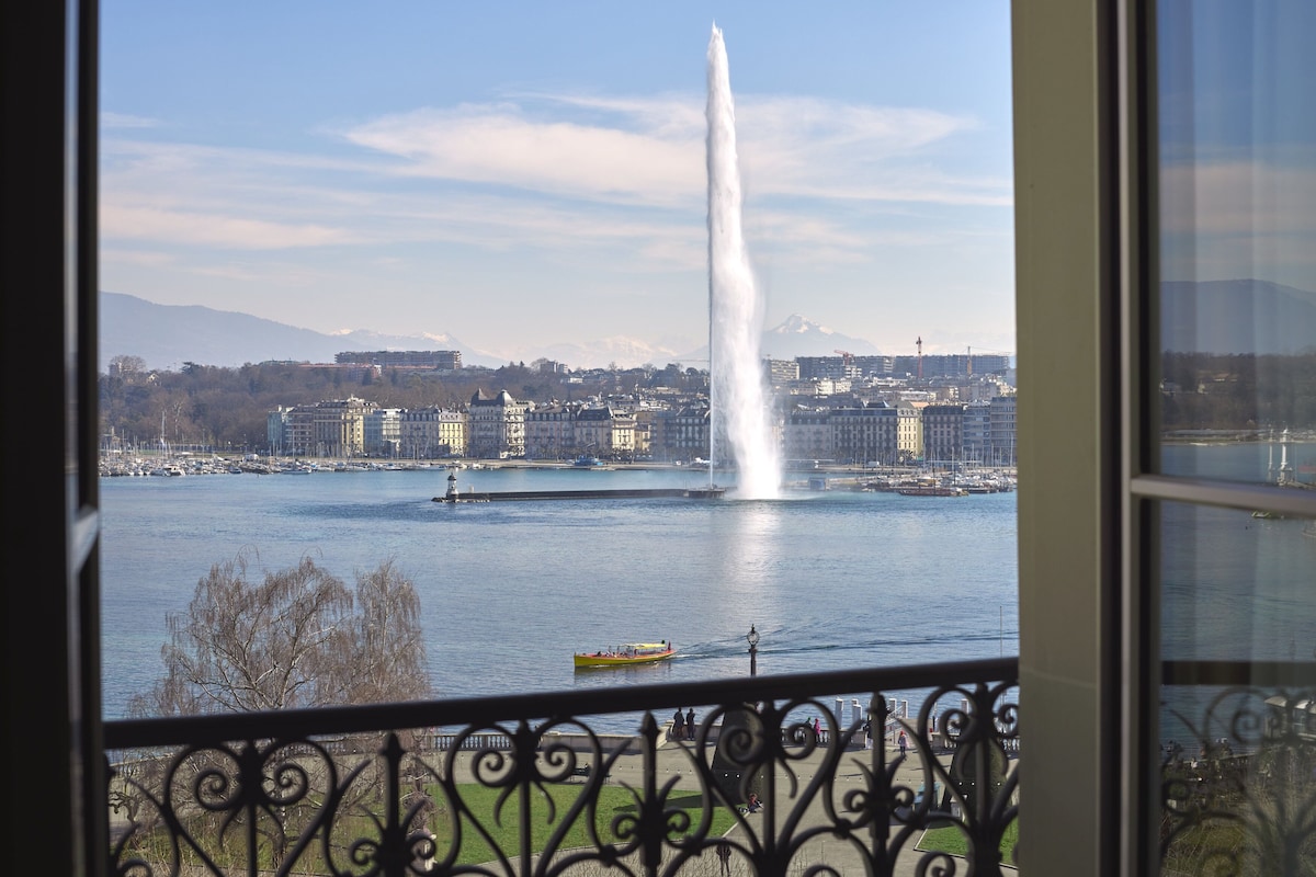 Une vue d’une ville avec une fontaine imposante au centre, des bâtiments en arrière-plan et un bateau sur l’eau.
