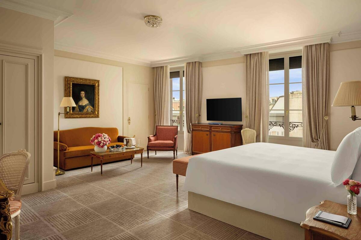 A hotel room with a large bed, a sofa, a desk with a lamp, a television, and a window with curtains.
