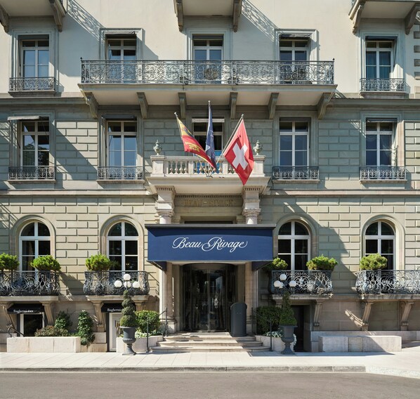 A hotel facade with a prominent entrance, flags, and a sign reading 'Beau Rivage.'