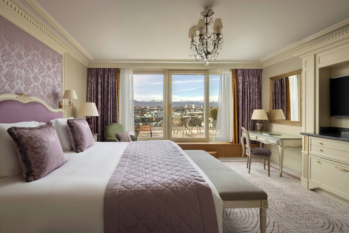 A hotel room with a large bed, a desk, a chair, a television, and a view of the cityscape.