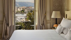 A hotel room with a bed, a nightstand with a lamp, and a view of a cityscape through a balcony. - Beau Rivage Geneva (Geneva)