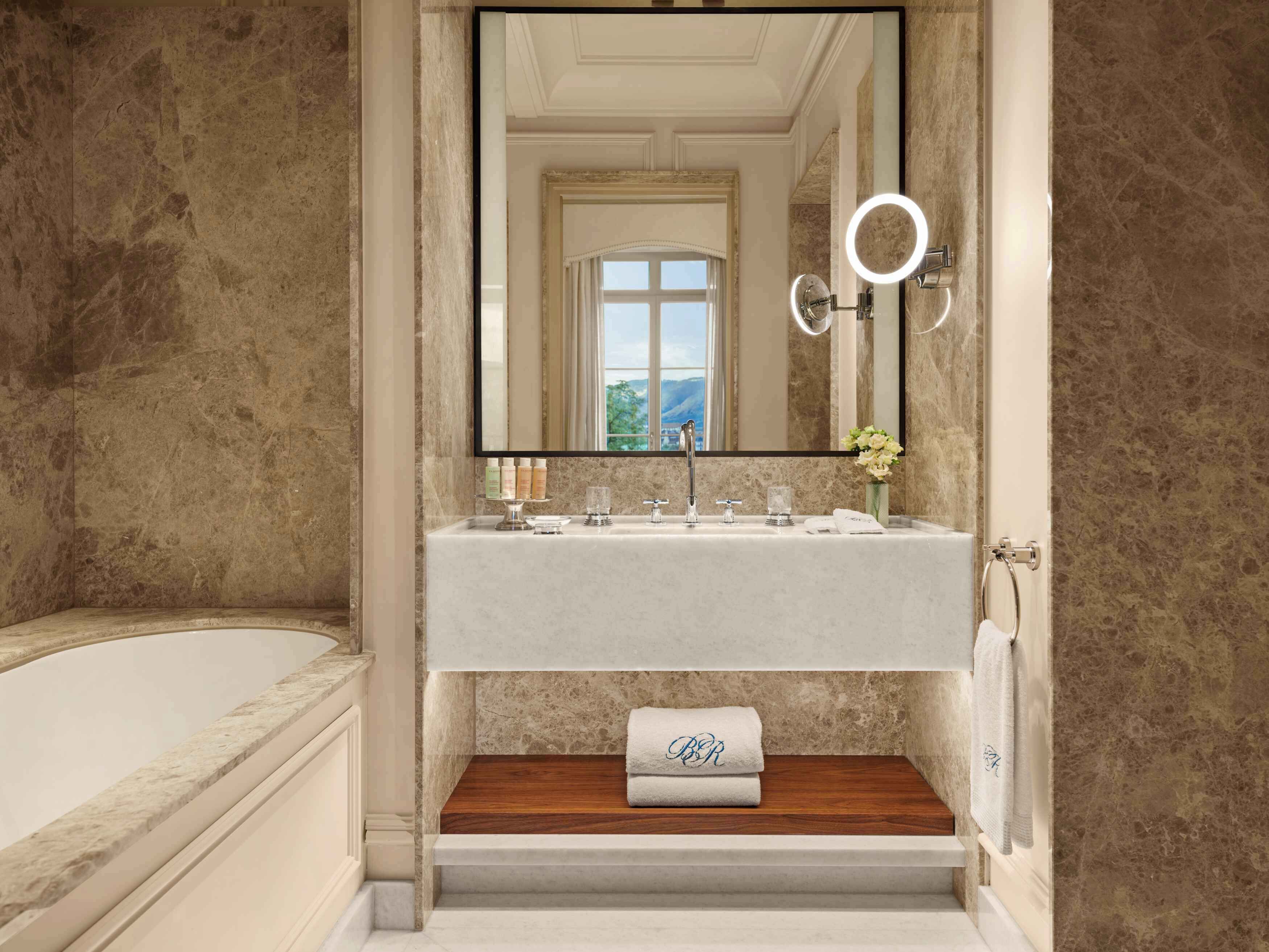 A bathroom with a marble wall, a large mirror, a sink with toiletries, and a towel with initials on a wooden shelf.