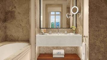 Baño con pared de mármol, un espejo grande, lavamanos con artículos de aseo y una toalla con iniciales sobre un estante de madera.