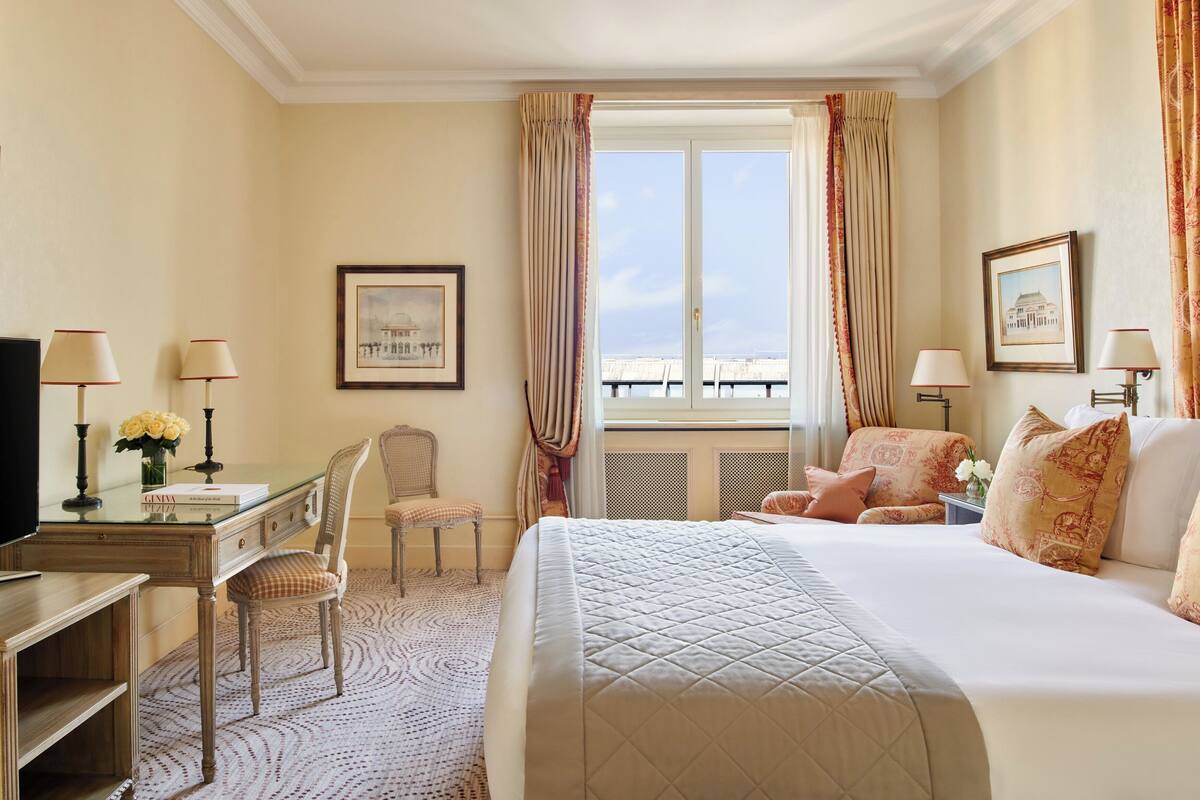 A hotel room with a bed, desk, chair, and a view of the sea through the window.