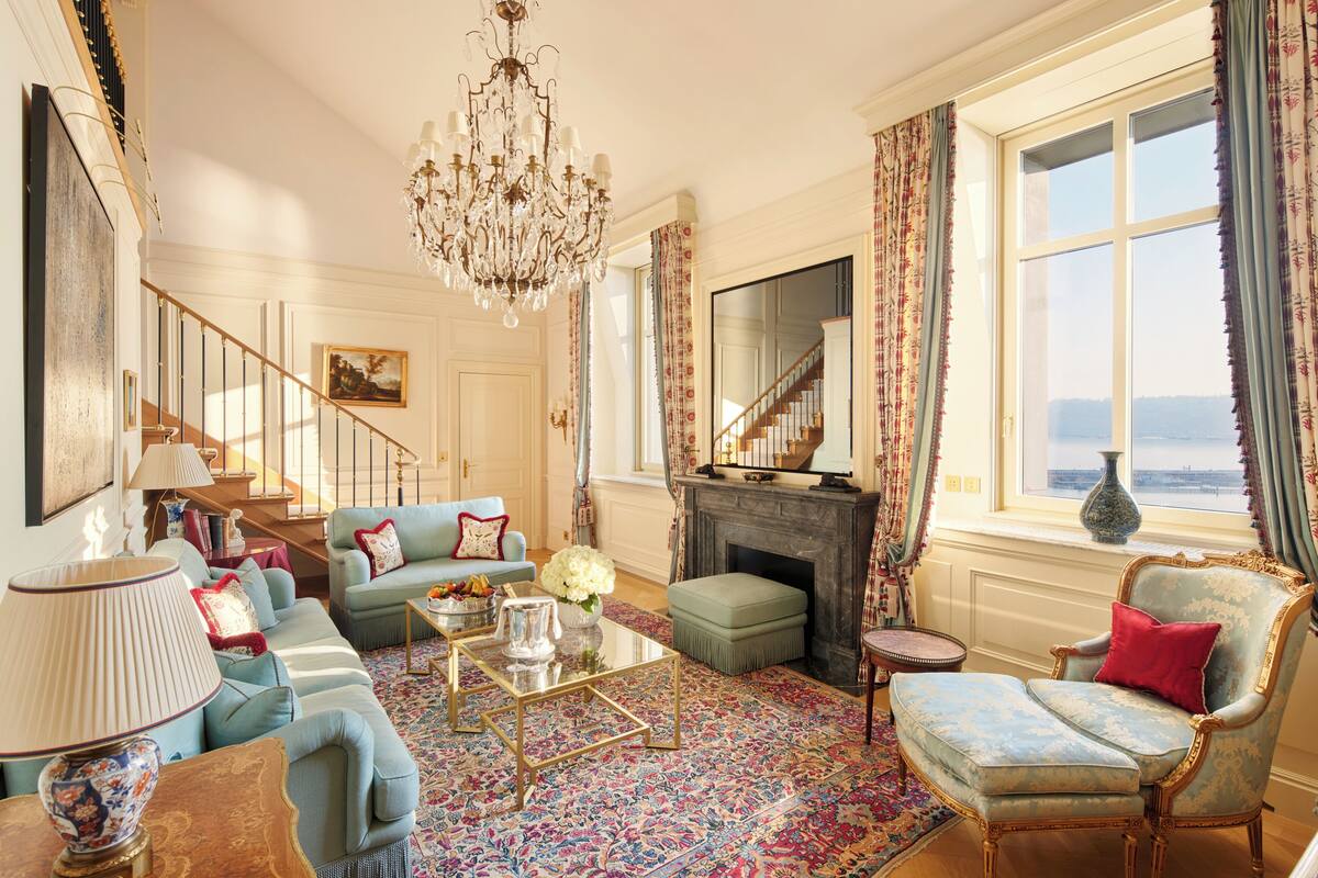 A spacious living room with a chandelier, a fireplace, a sofa set, and a view through a large window.