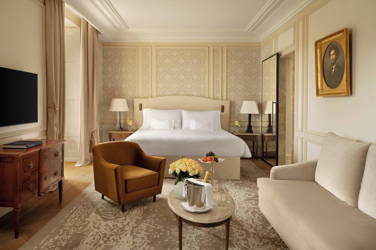 A hotel room with a large bed, two bedside tables, a TV, a sofa, a chair, and a small table with flowers and a champagne bottle.