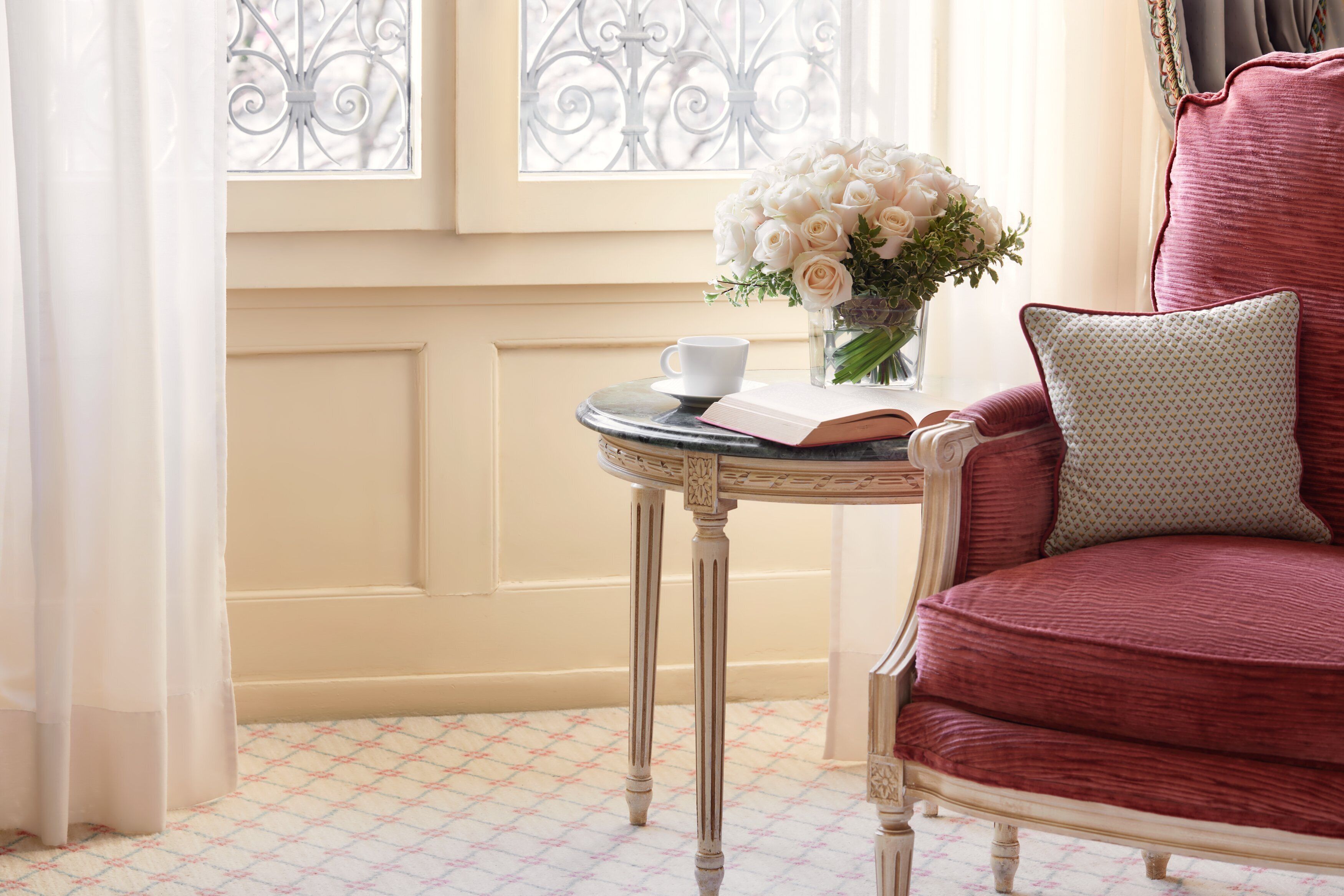 Una habitación con una butaca tapizada en rojo, una mesita redonda con un libro y una taza, y un florero junto a una ventana con rejas decorativas de hierro.