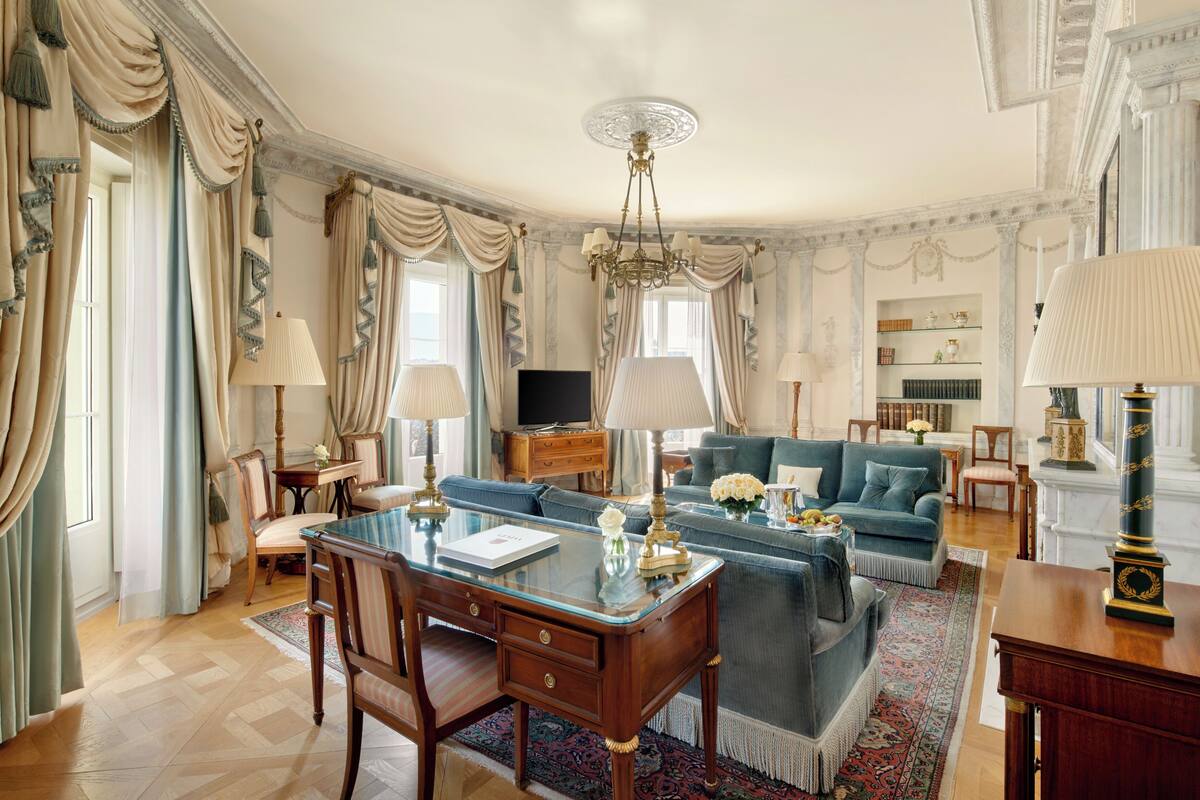 A spacious living room with a glass coffee table, a blue sofa, and a wooden desk with a lamp.