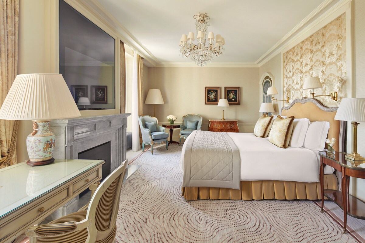 A spacious bedroom with a large bed, a desk, a chair, a fireplace, and a chandelier.