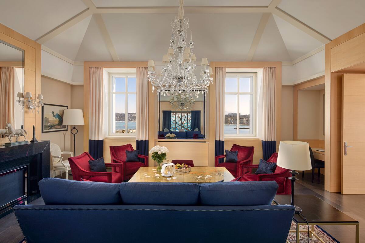 A spacious living room with a large blue sofa, red armchairs, a chandelier, and a view of the city through large windows.
