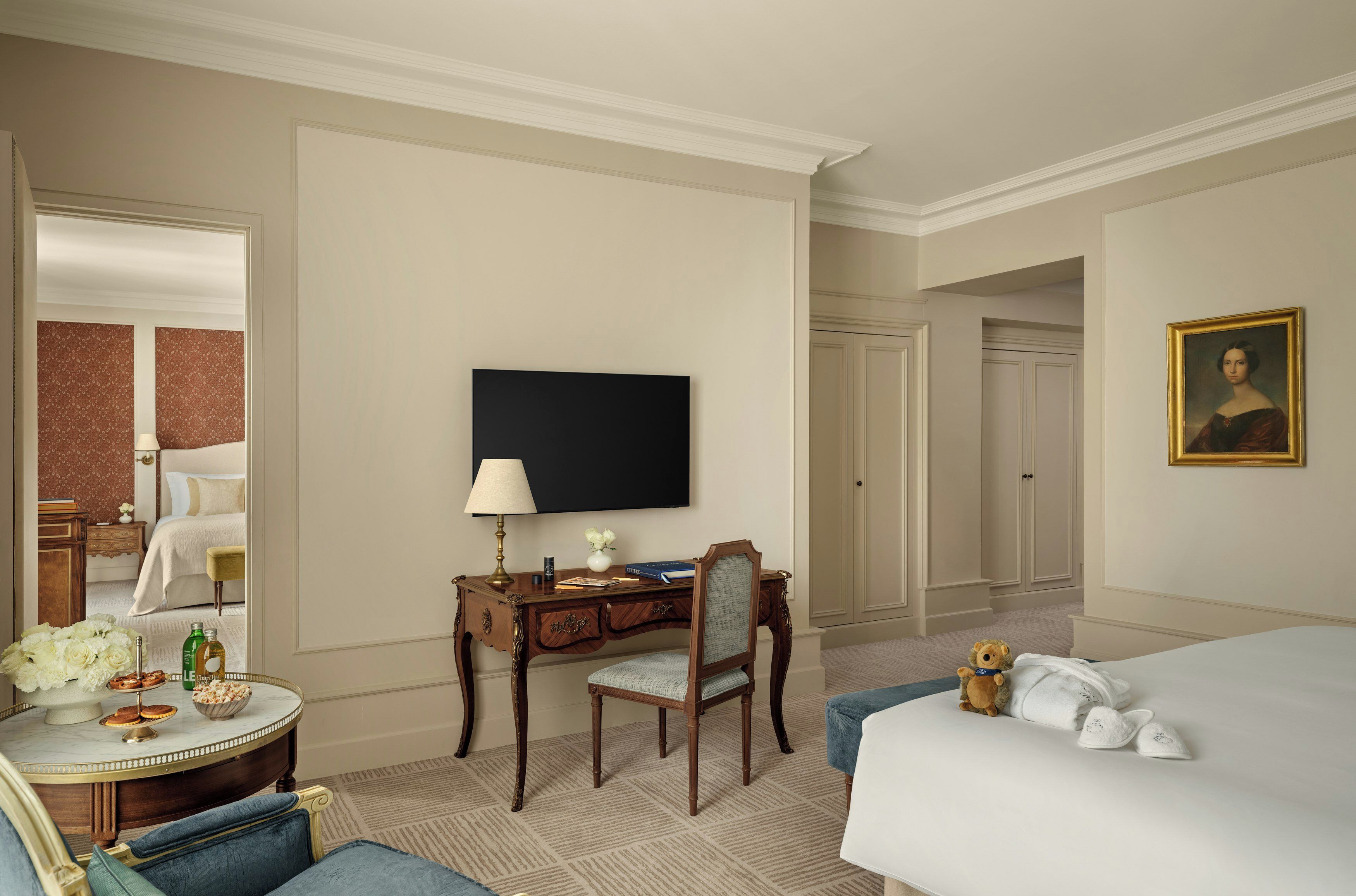 A hotel room with a bed, a desk with a lamp, a television, a painting, and a teddy bear.