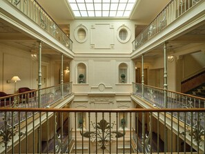 A grand interior with a central atrium, ornate railings, and a staircase leading to an upper level.