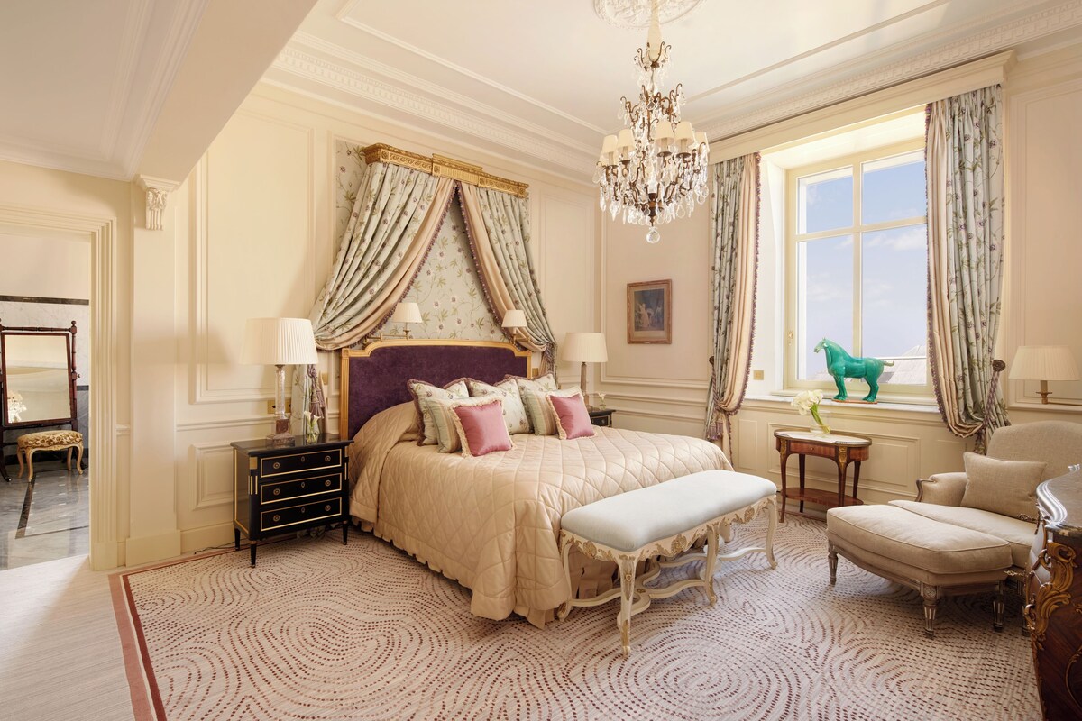 A luxurious bedroom with a large bed, ornate curtains, a chandelier, and a view of the outdoors.