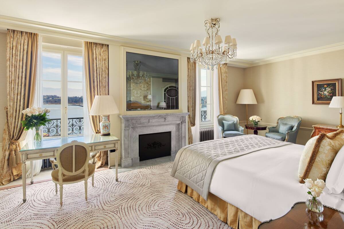 A spacious bedroom with a large bed, a sitting area with a chair, a desk, and a fireplace.