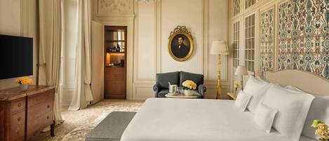 A luxurious bedroom with a large bed, a television, a desk with drawers, a chair, and a chandelier.