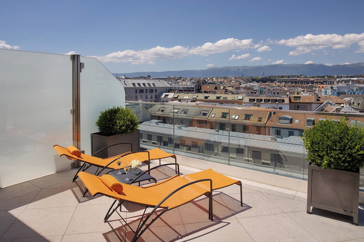 A rooftop terrace with lounge chairs, a glass balustrade, and a cityscape view.
