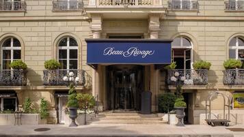 L’entrée d’un hôtel, surmontée d’une enseigne indiquant « Beau Rivage », se caractérise par un portail imposant, des balcons richement décorés et des plantes en pot.
