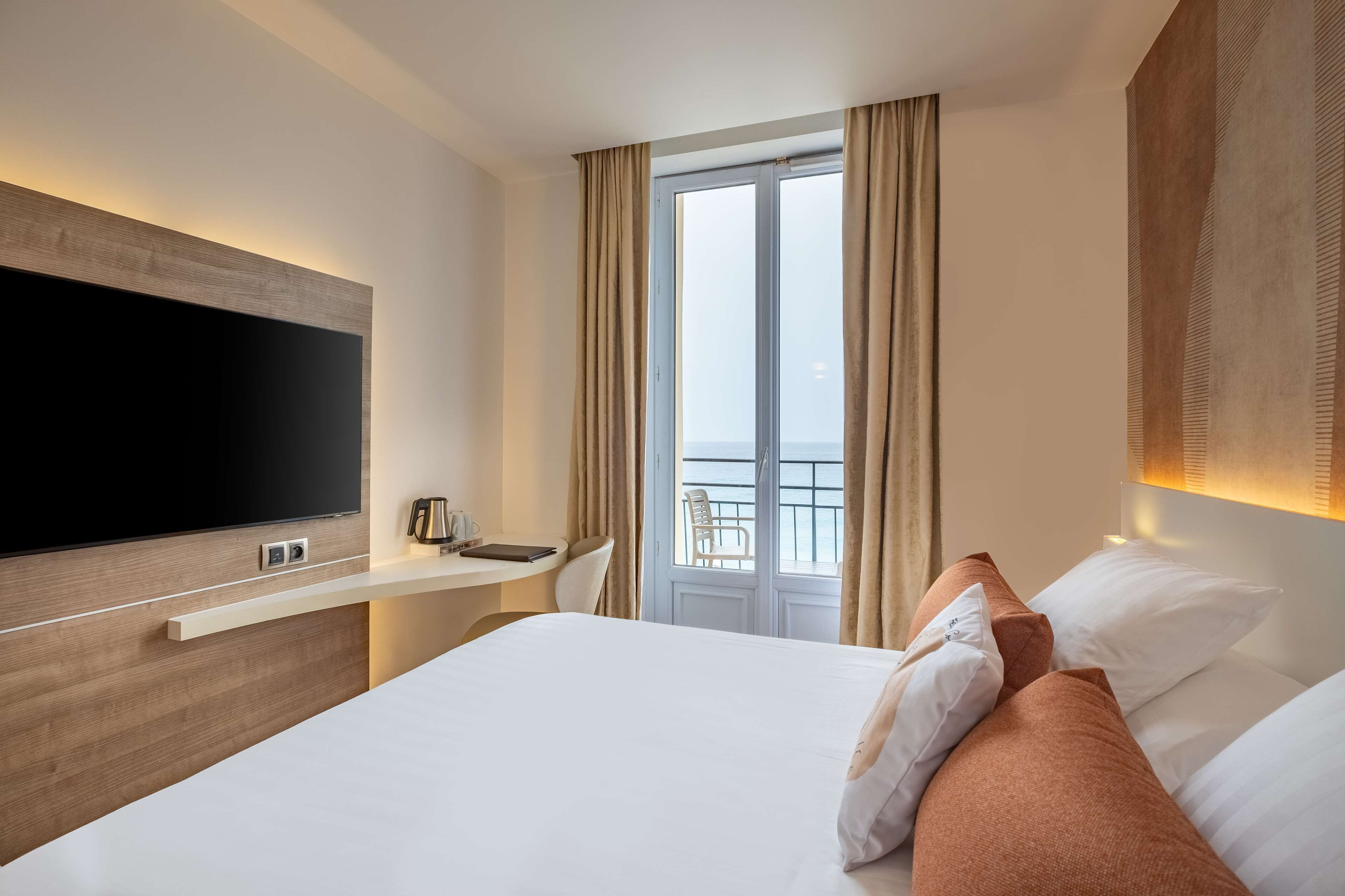 Ett hotellrum med en stor säng, en platt-TV, ett skrivbord med en stol och en balkong med gardiner.