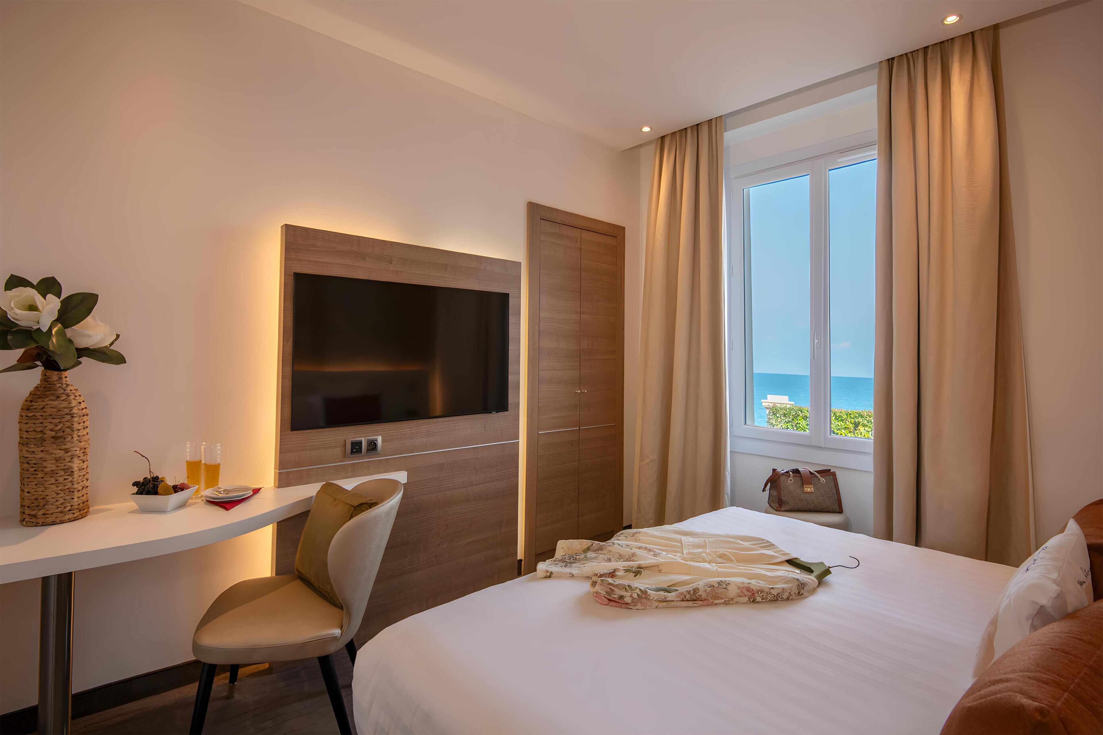 Habitación de hotel con cama, televisor, escritorio con un florero y vasos, silla y vista al mar a través de una ventana.