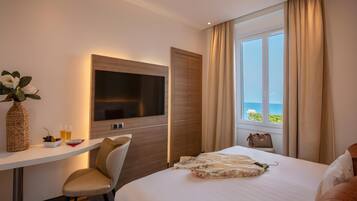 Habitación de hotel con cama, televisor, escritorio con un florero y vasos, silla y vista al mar a través de una ventana.