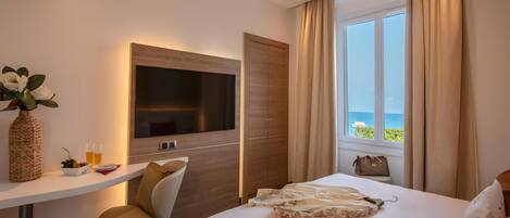 A hotel room with a bed, a TV, a desk with a vase and glasses, a chair, and a view of the sea through a window.
