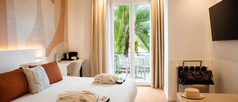 A hotel room with a bed, a desk, a chair, a TV, and a balcony with a view of greenery.