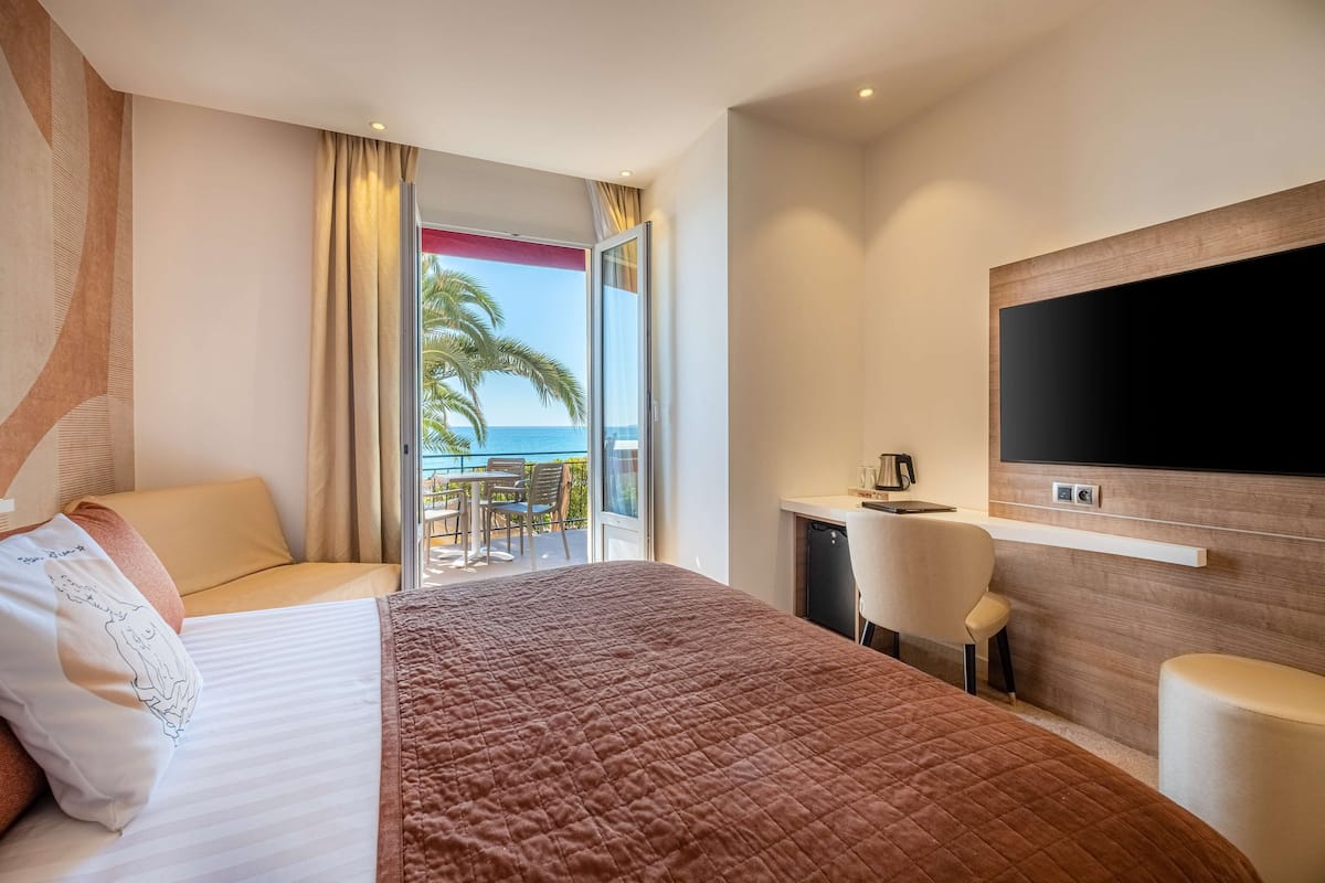 A hotel room with a bed, a desk, a chair, a TV, and a balcony with a view of the ocean.