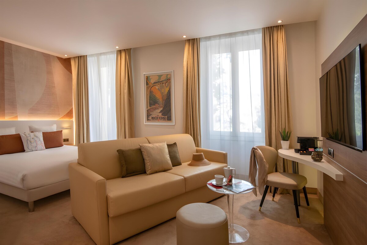 A modern hotel room with a bed, sofa, desk, chair, and a large TV.