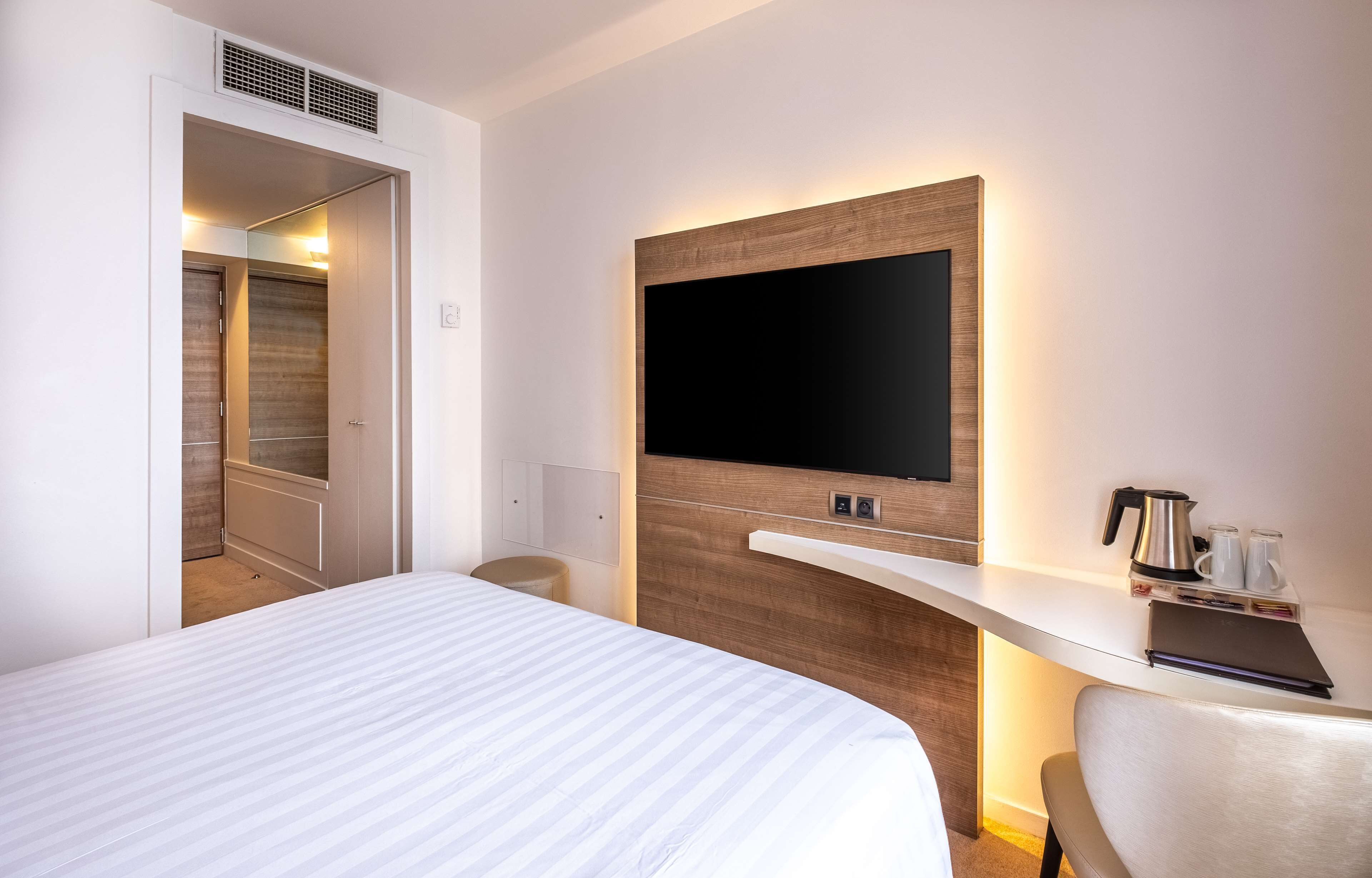 Una habitación de hotel con una cama grande, un televisor de pantalla plana montado en un panel de madera, un escritorio pequeño con una tetera y tazas, y una silla.