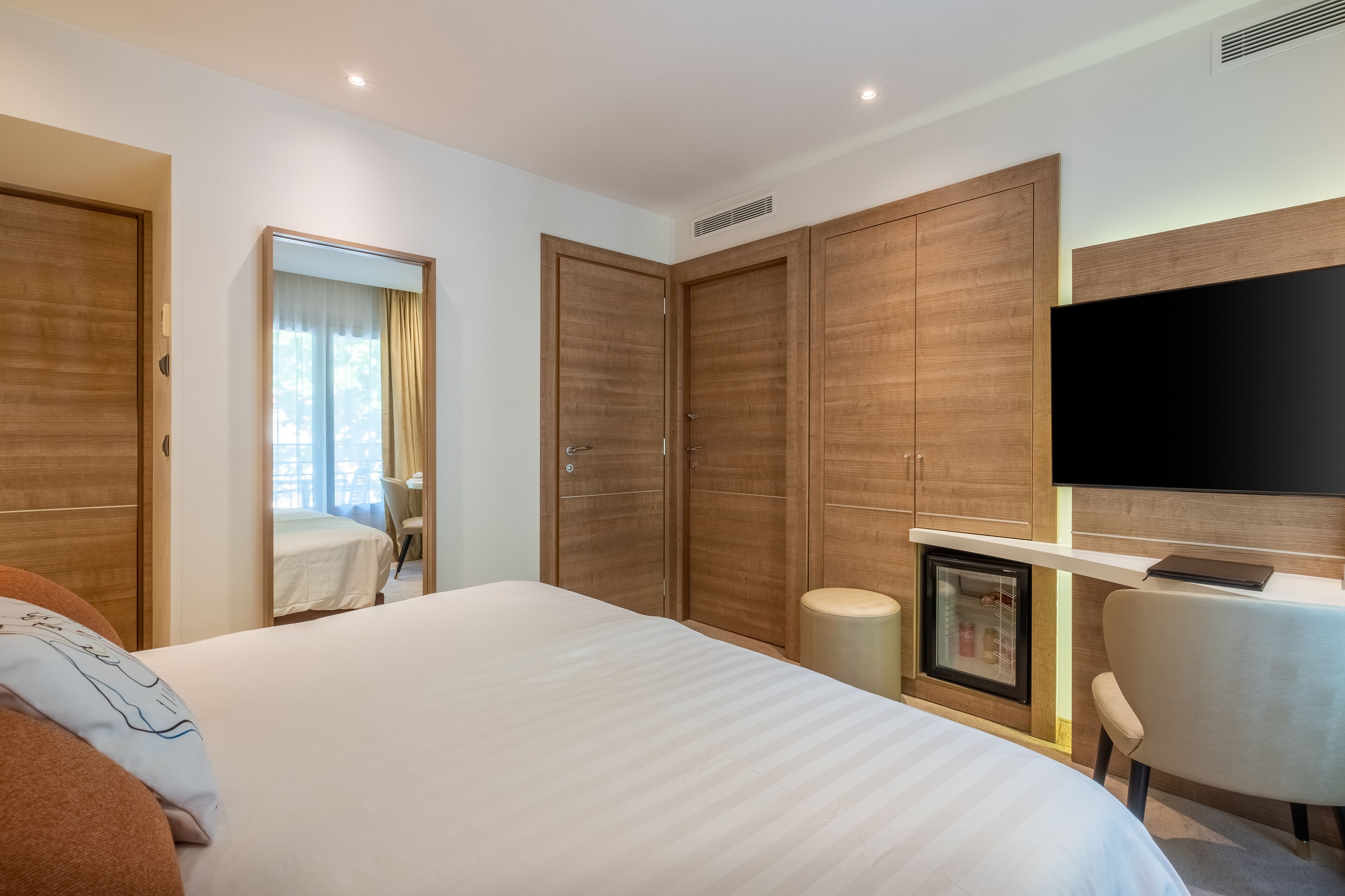 A modern hotel room with a large bed, wooden wardrobes, a desk with a chair, a television, and a small refrigerator.