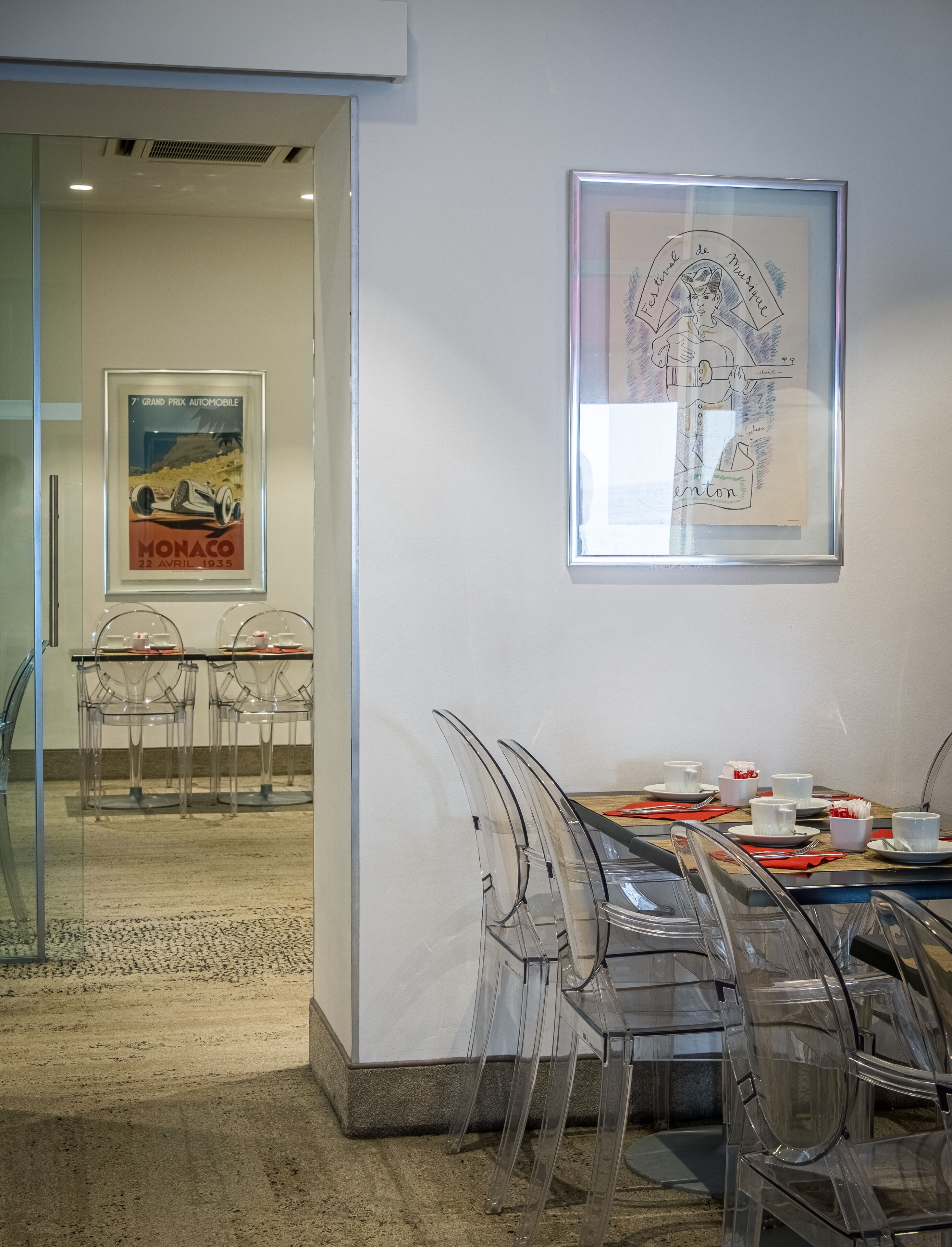 Un salón de conferencias con un separador de vidrio, sillas de plástico transparente, una mesa de madera dispuesta con tazas y platos blancos, y dos obras de arte enmarcadas en la pared.