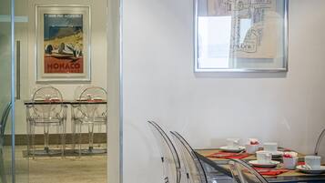 Un salón de conferencias con un separador de vidrio, sillas de plástico transparente, una mesa de madera dispuesta con tazas y platos blancos, y dos obras de arte enmarcadas en la pared.