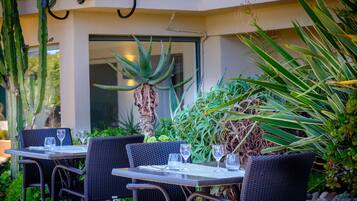 Uma área de refeições ao ar livre com cadeiras e mesas de vime, cercada por diversas plantas e uma grande planta de aloe vera.