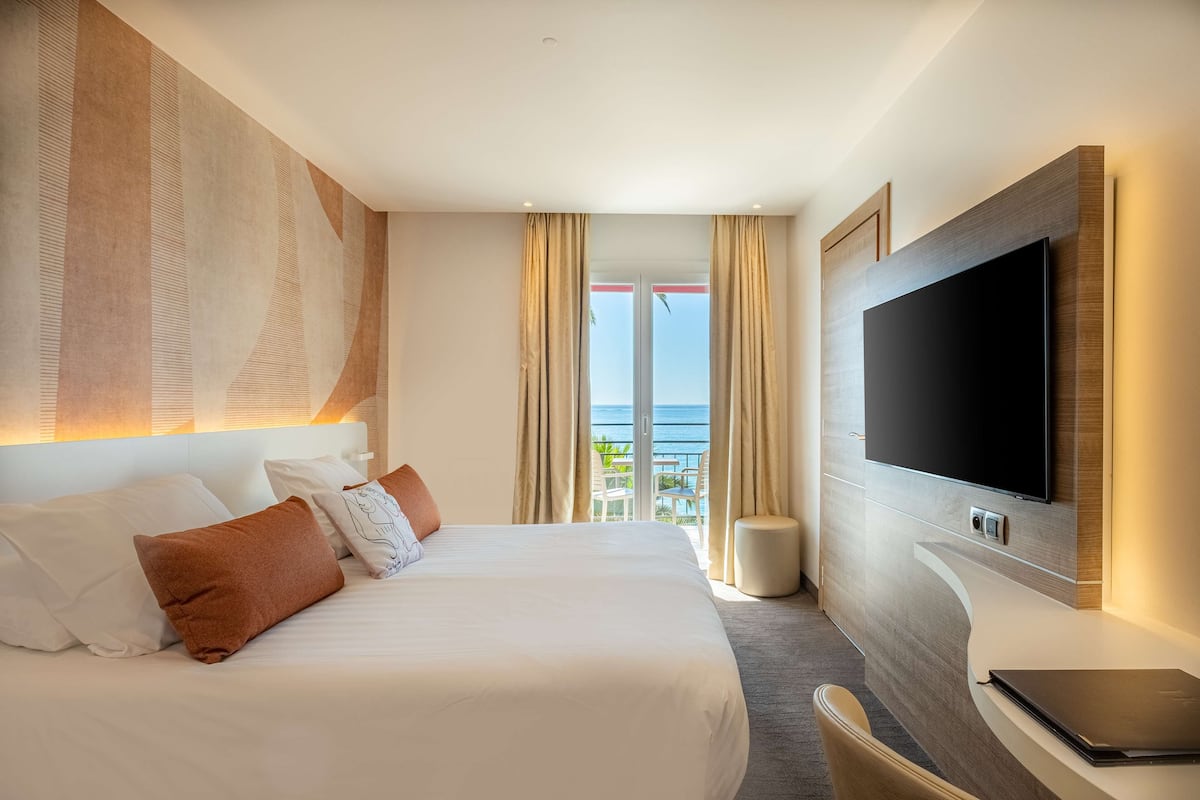 A hotel room with a large bed, a flat-screen TV, a desk with a laptop, and a view of the ocean through the window.