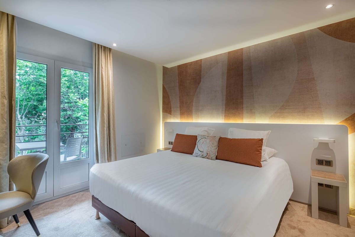 A modern hotel room with a large bed, a chair, a nightstand, and a view of greenery through the window.