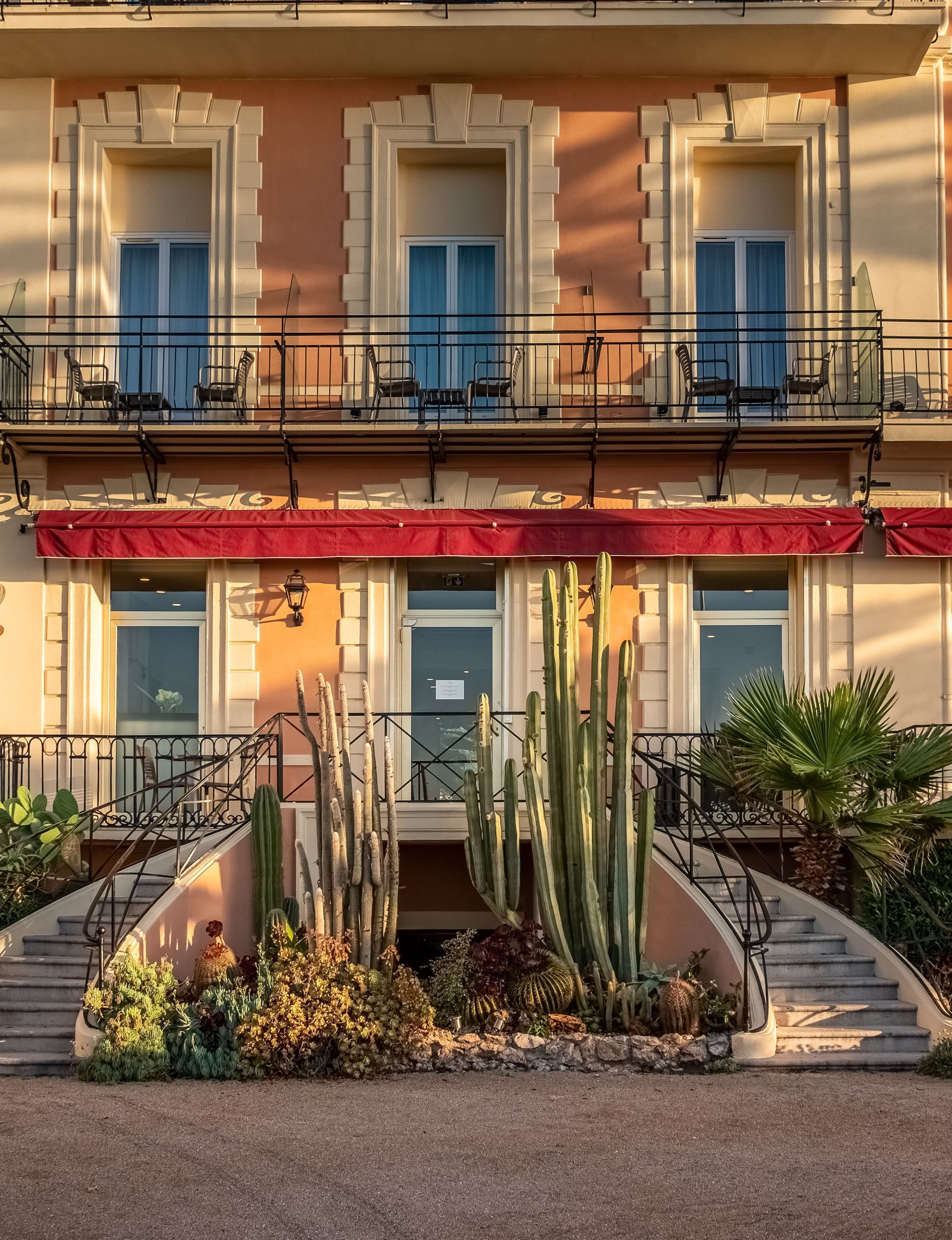 A two-story building with a red awning, balconies with chairs, and a cactus garden in front.