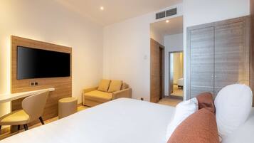 A modern hotel room with a large bed, a flat-screen TV mounted on the wall, a desk with a chair, a sofa, and a wardrobe.