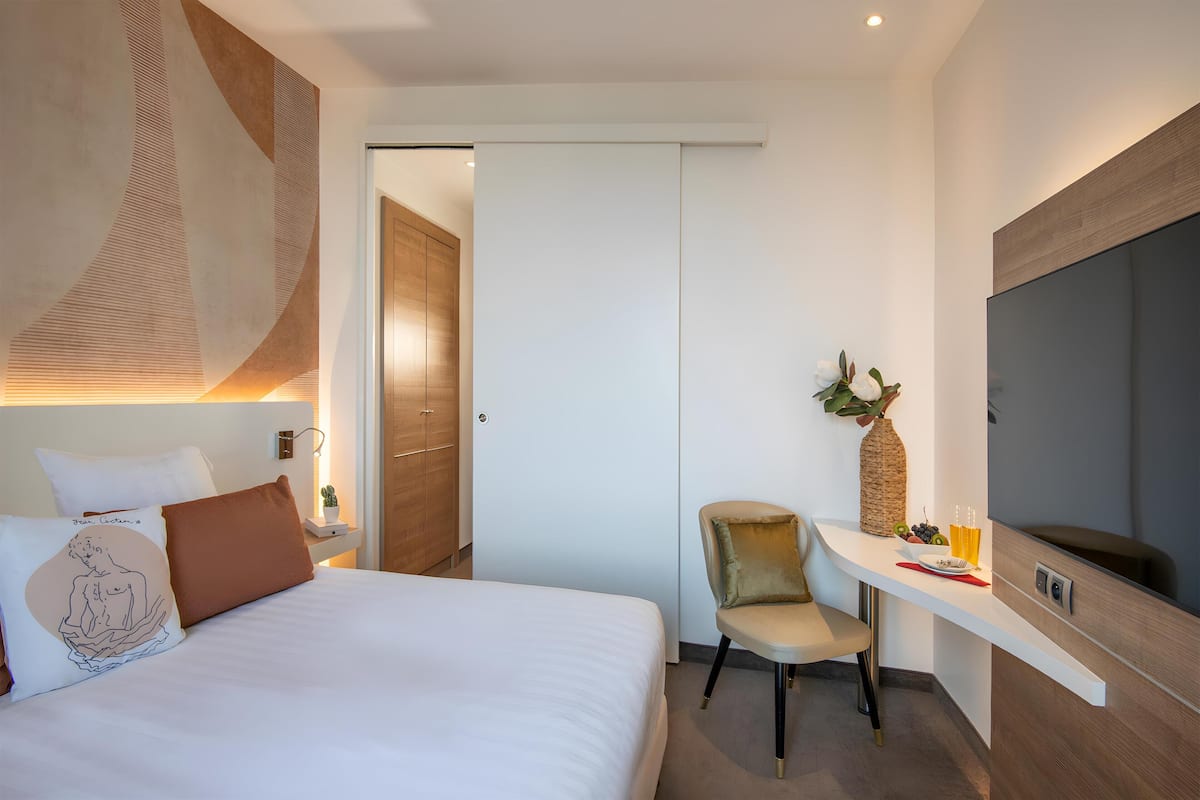 A modern hotel room with a bed, bedside table, chair, TV, and a door leading to another room.