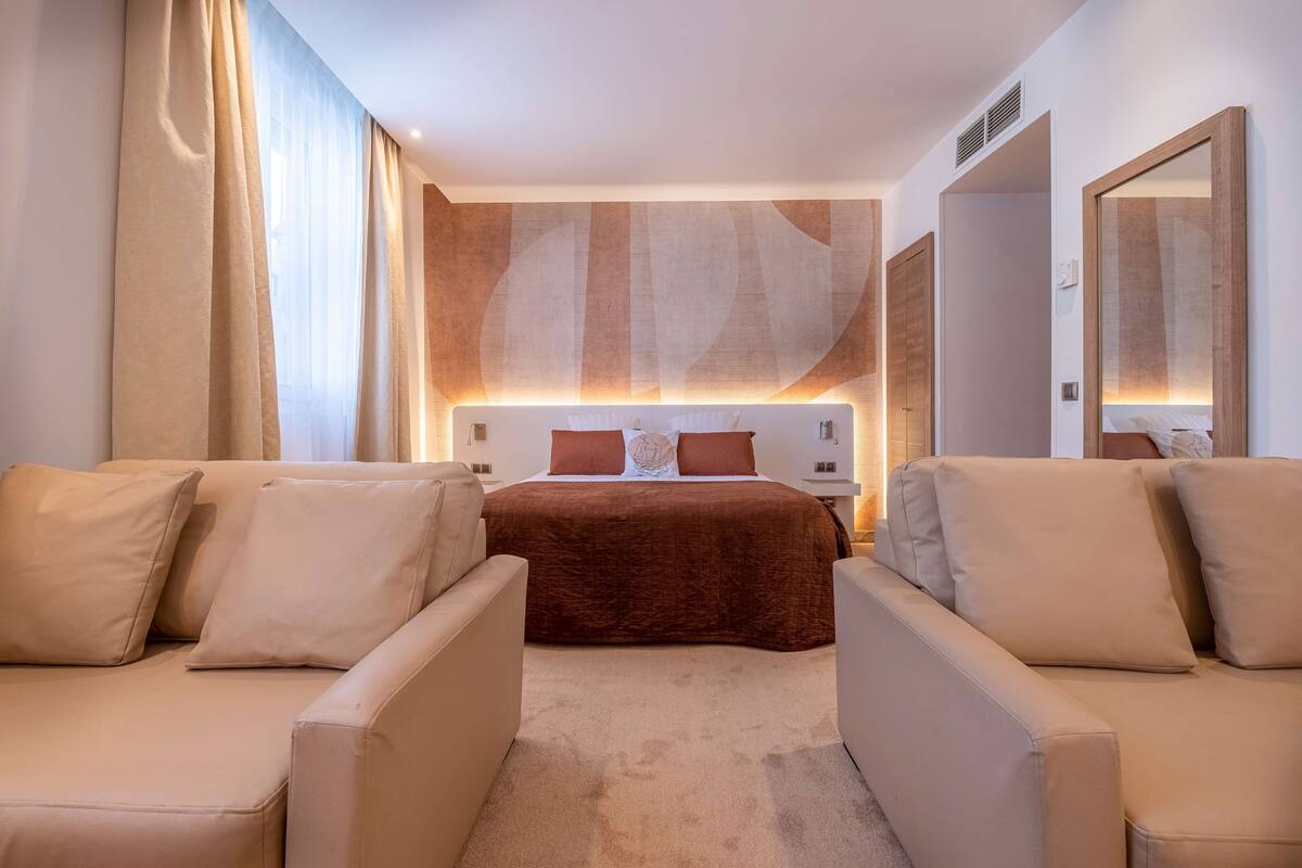 A modern hotel room with a large bed, two armchairs, a nightstand, and a large mirror.