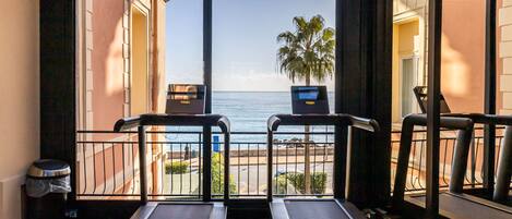 A room with two treadmills facing a balcony with a view of the sea and palm trees.