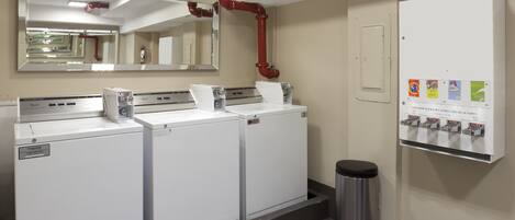 A laundry facility with three white washers and dryers, a trash bin, a mirror, and a dispenser with beverage options.