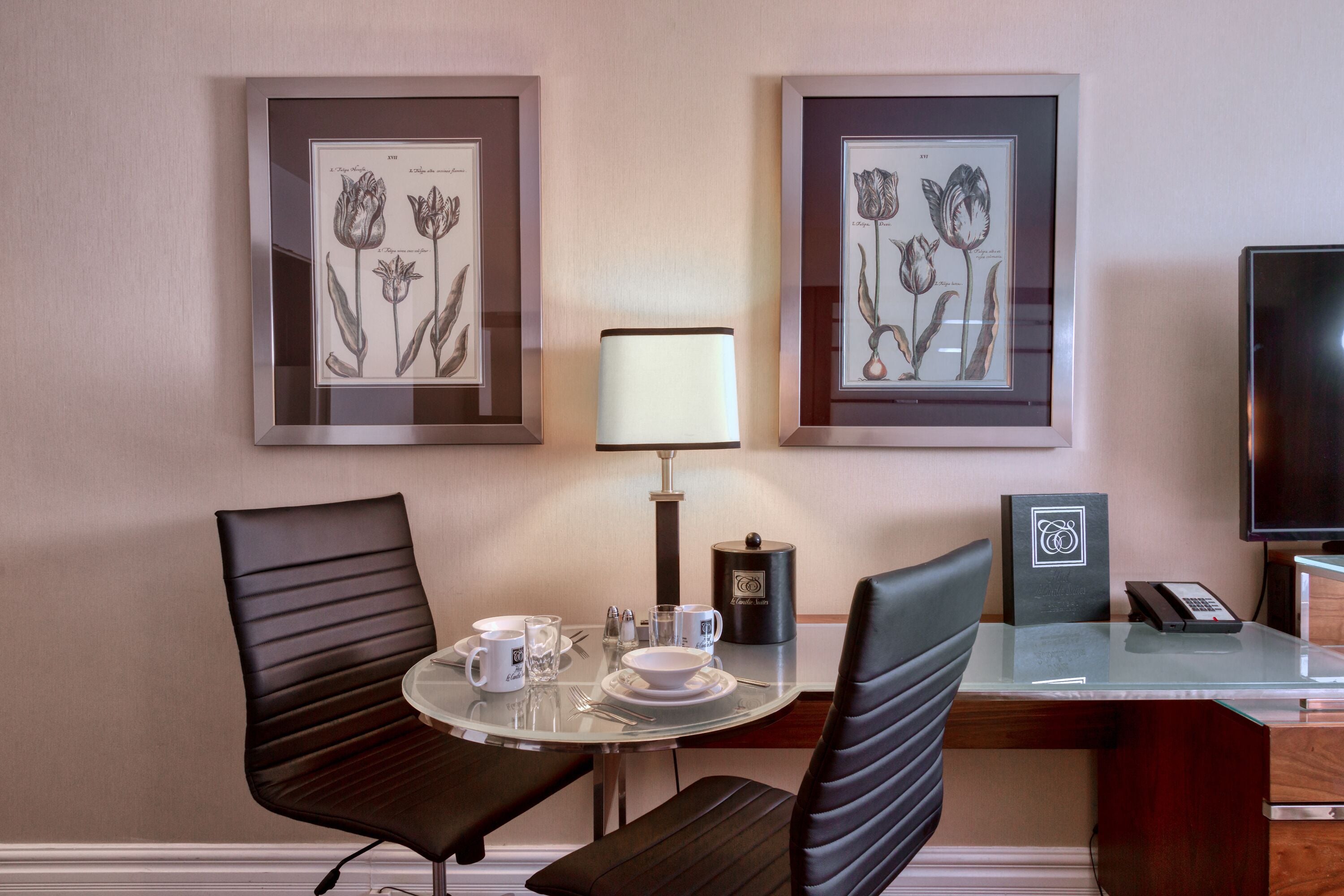 A hotel room with a dining area featuring two framed botanical prints, a table with a lamp, chairs, and a television.