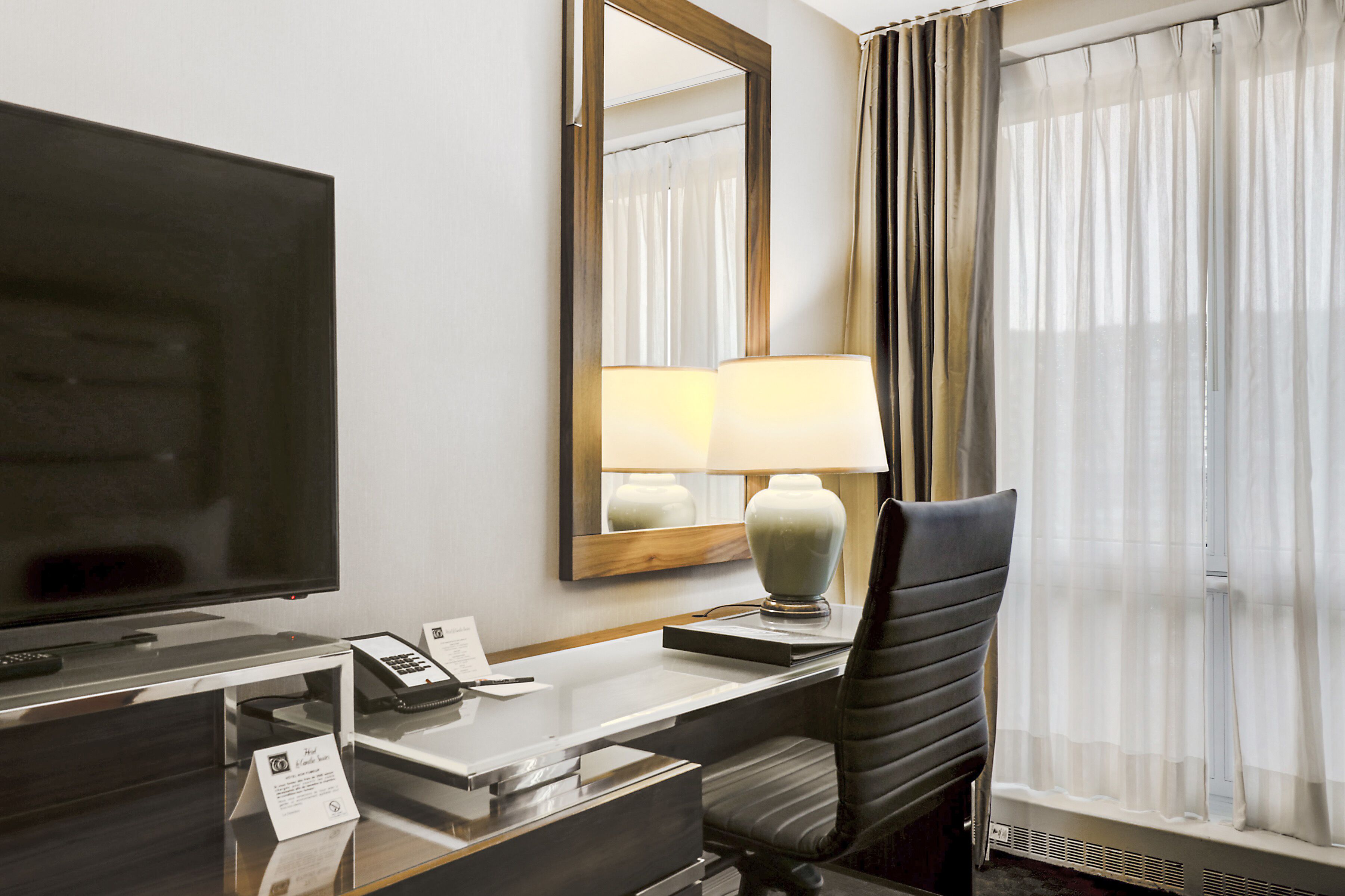 A hotel room with a flat-screen TV, a desk with a lamp, a chair, and a large mirror.