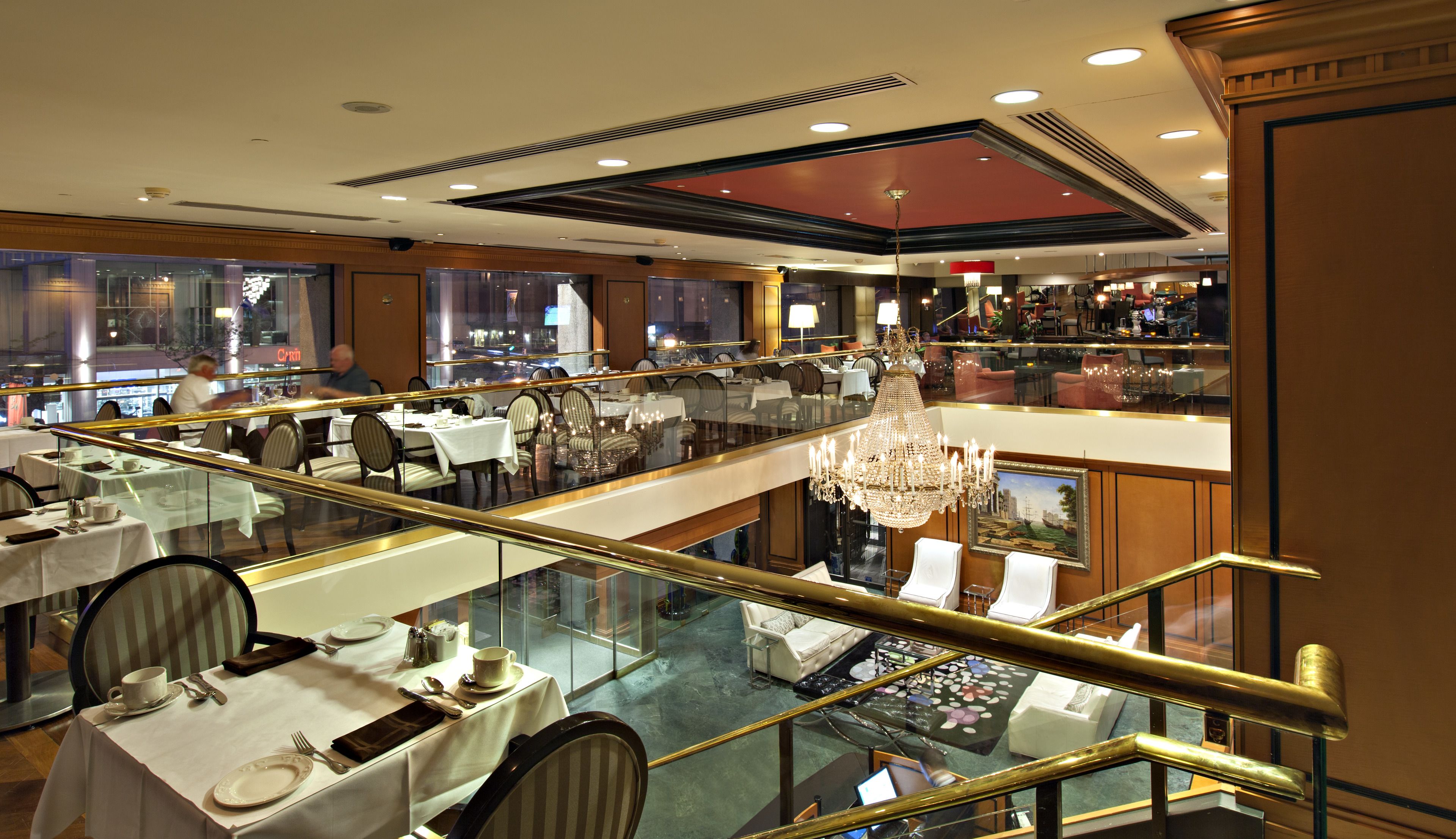 A multi-level dining area with glass railings, chandeliers, and a view of the cityscape.