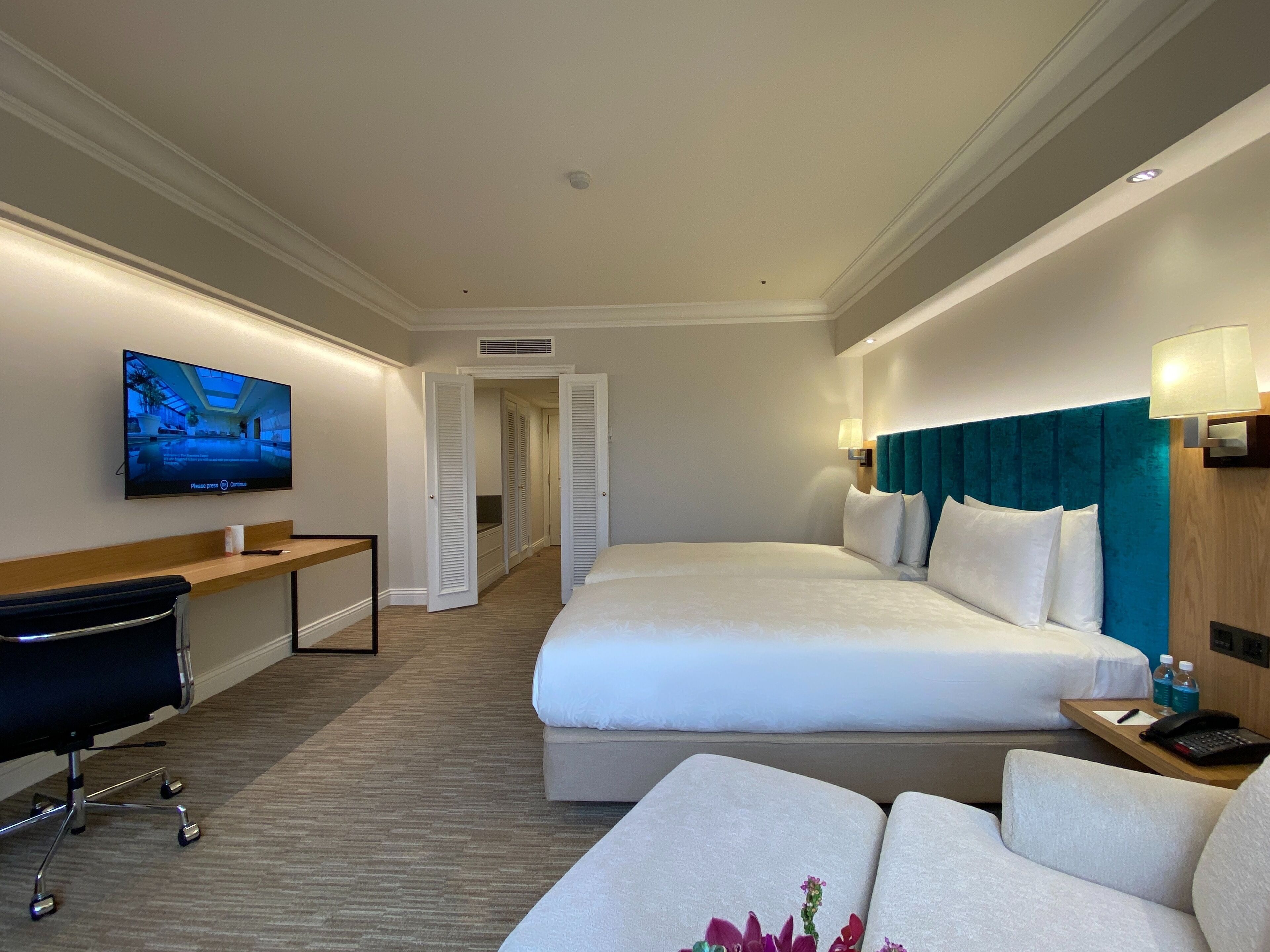 A hotel room with a bed, a desk, a chair, a TV, and a bathroom visible through an open door.