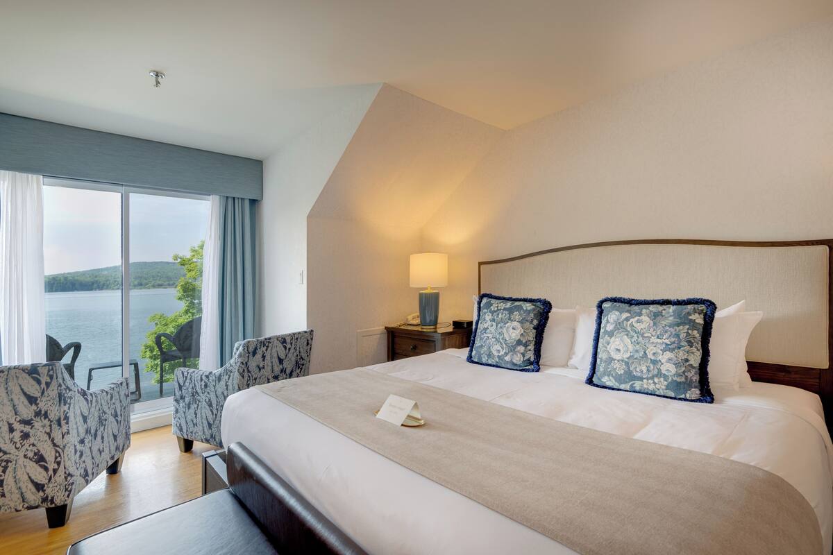 A hotel room with a large bed, two matching chairs, a nightstand with a lamp, and a view of the water.