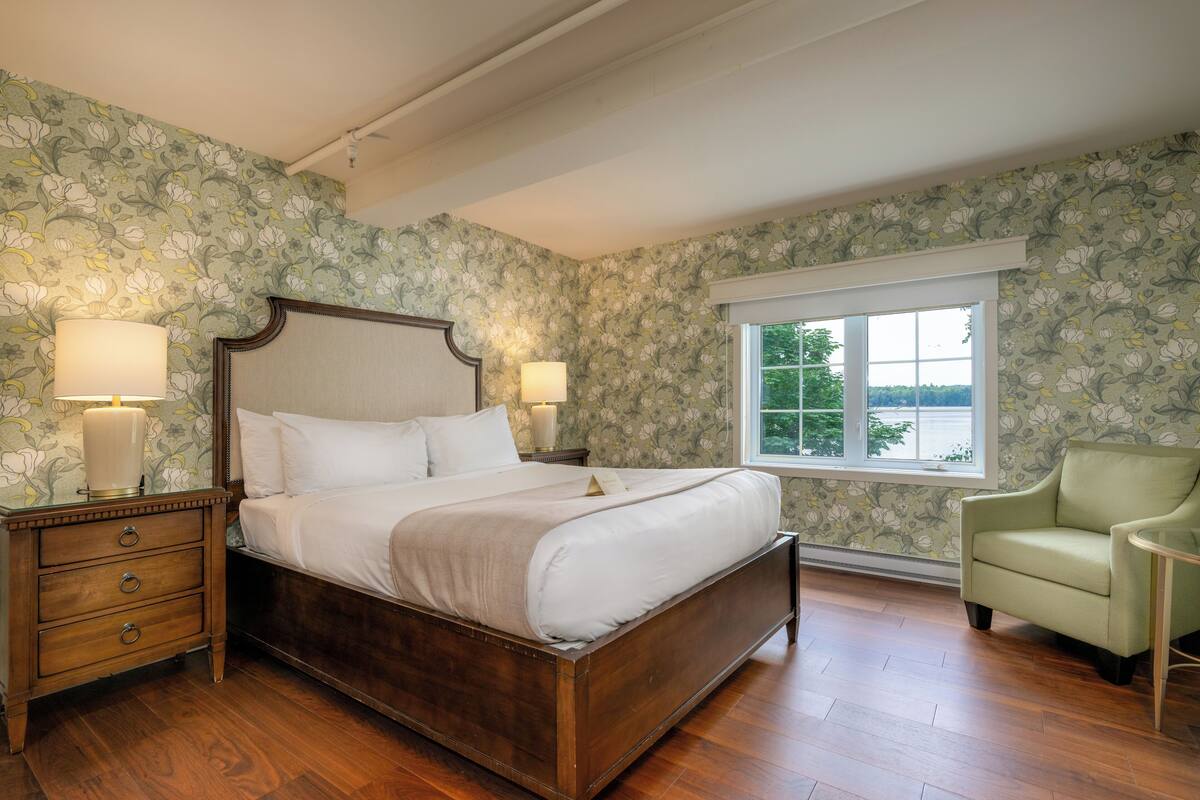 A bedroom with a wooden bed, two bedside lamps, a nightstand, a green armchair, and a window with a view of trees.