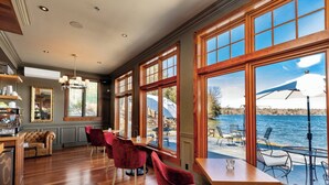 A dining area with wooden tables and red chairs, large windows with a view of a body of water, and a chandelier.